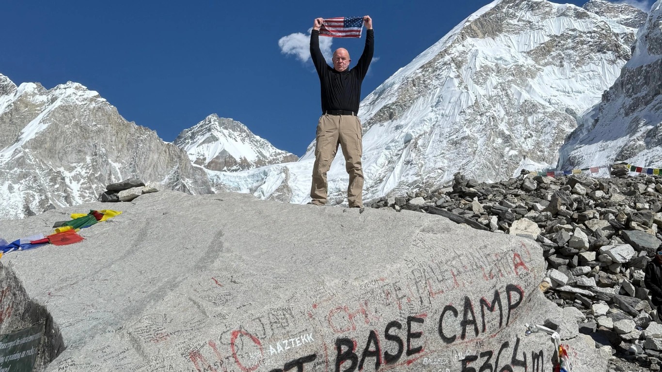 Older American trekker proudly holding the U.S. flag on the famous Everest Base Camp stone during the Everest Base Camp Trek in Nepal.