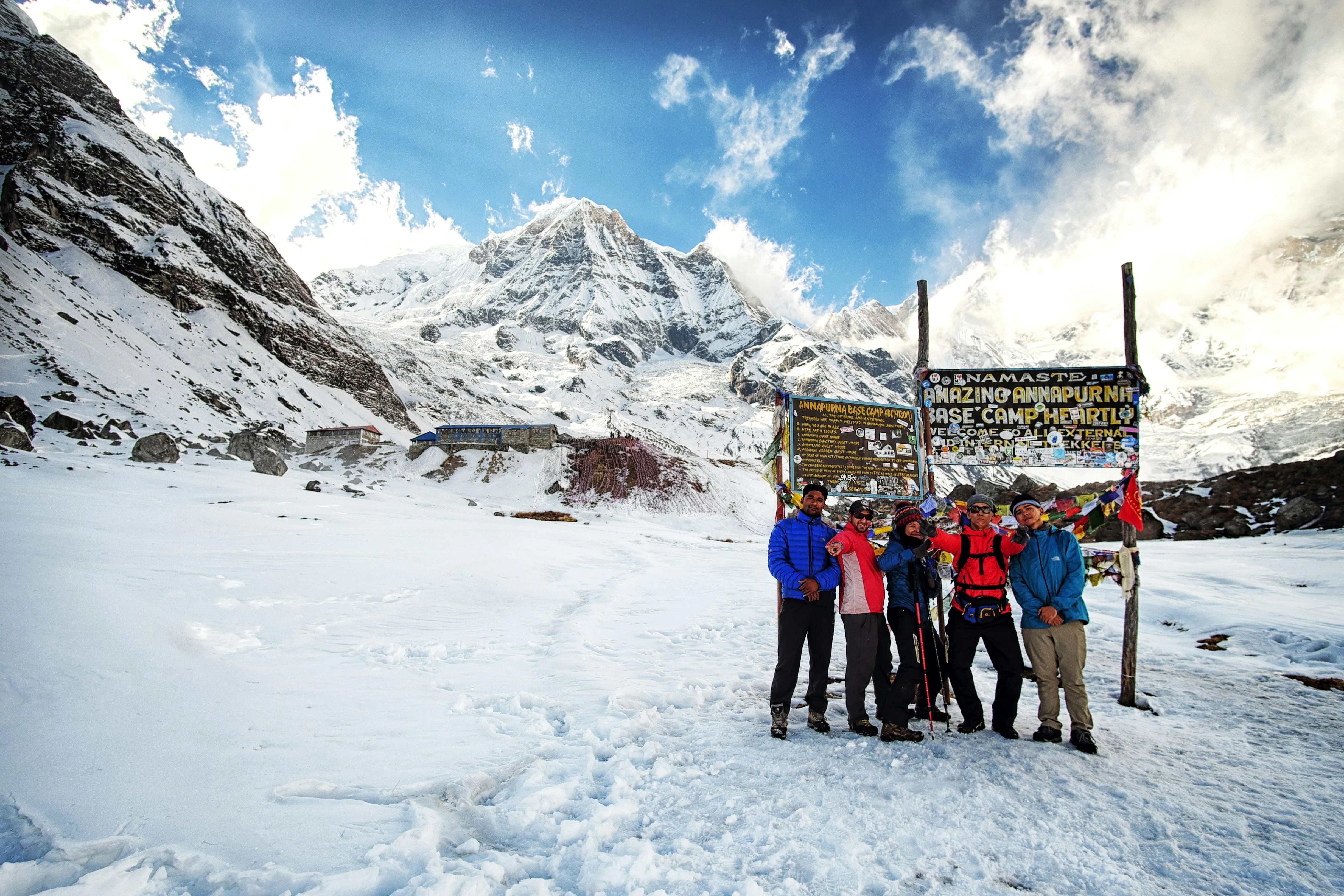 Trekking in Nepal at Annapurna Base Camp, with hikers posing in front of the signboard and towering Himalayan peaks behind them.