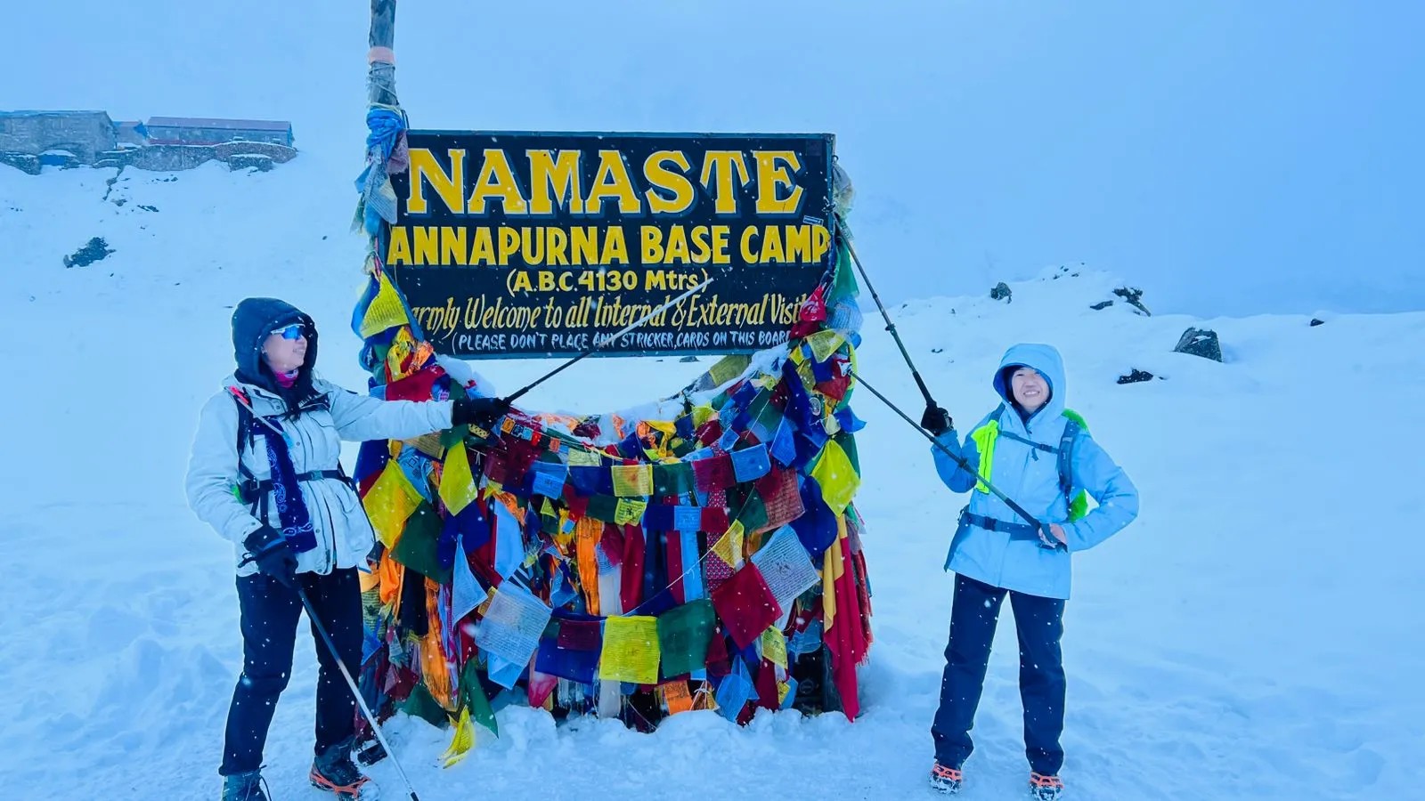 Two female trekkers posing at the Annapurna Base Camp signboard during heavy snowfall, showcasing the Annapurna Base Camp trek difficulty in harsh winter conditions and high-altitude weather.