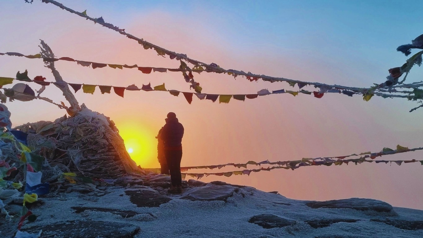 Trekker at the summit of Pikey Peak Trek Nepal with vibrant prayer flags fluttering and panoramic Himalayan mountain views in the background.