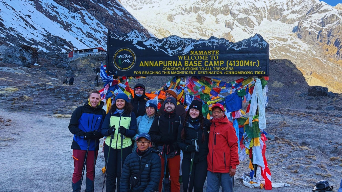 Group of trekkers and their guide taking a photo in front of the Annapurna Base Camp signboard at 4,130 meters surrounded by Himalayan peaks.