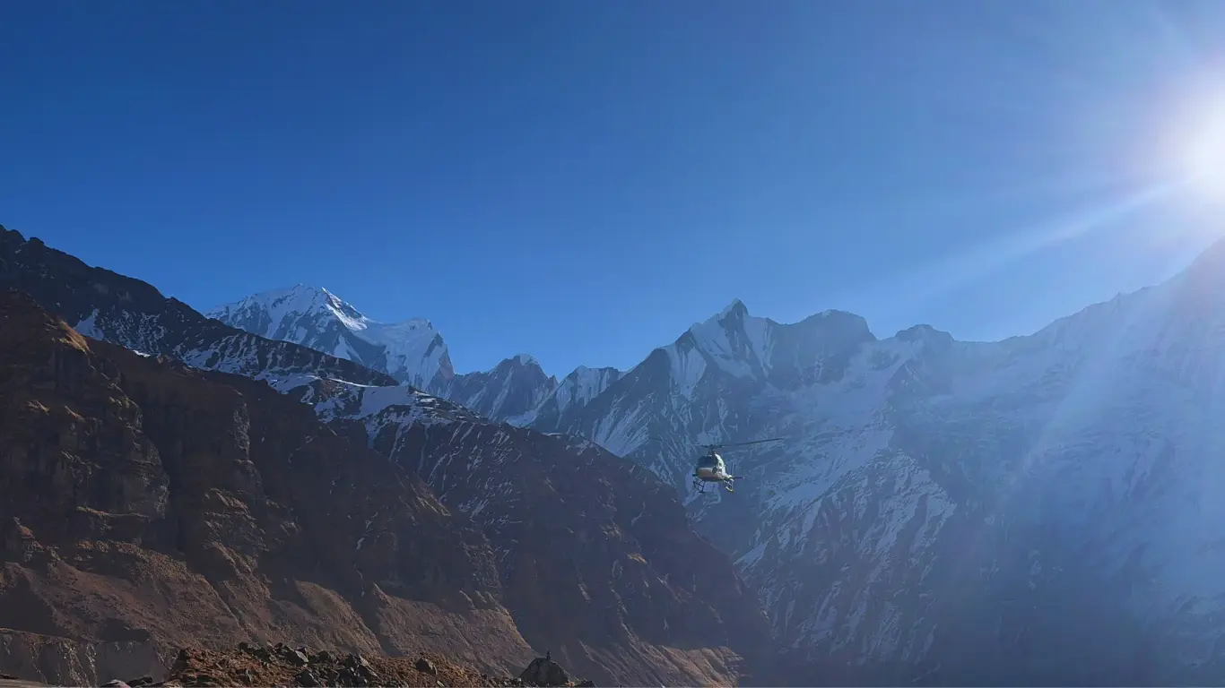 Helicopter flying towards Annapurna Base Camp with snow-capped peaks of the Annapurna range in the background, preparing to land.