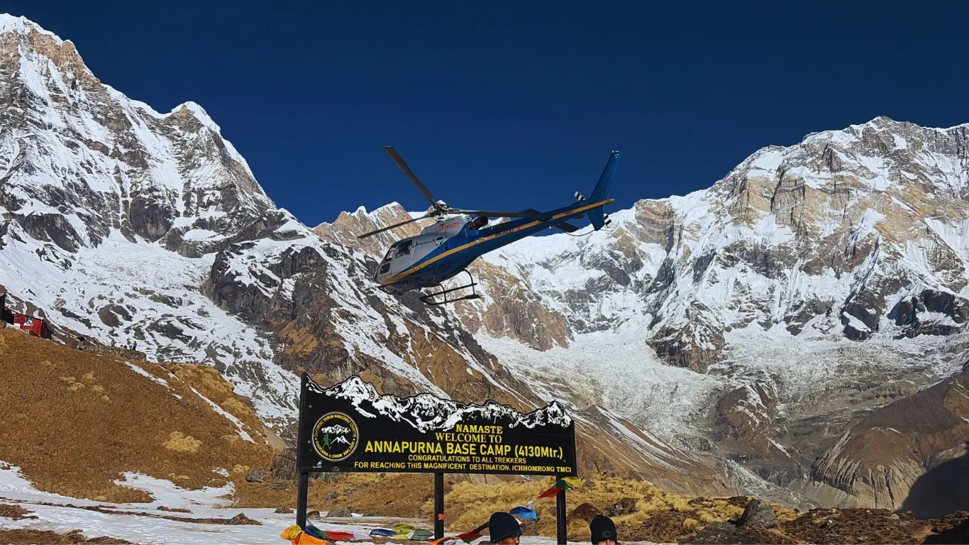 Helicopter flying above Annapurna Base Camp signboard with Annapurna I and Annapurna South in the background, preparing to land.