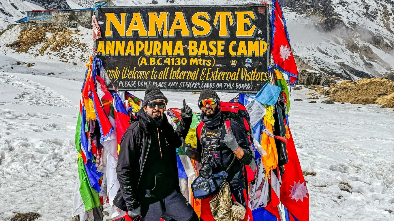 Two young trekkers smiling in front of the Annapurna Base Camp signboard.