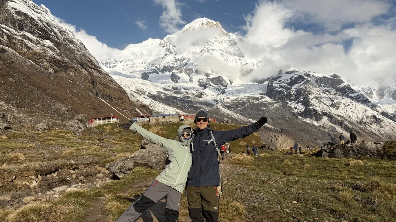 Two young trekkers smiling in front of the Annapurna Base Camp signboard.